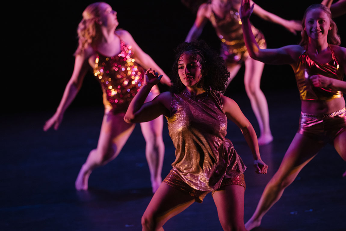 A college student in a gold dress dances on stage in dim lighting with a couple other dancers behind her